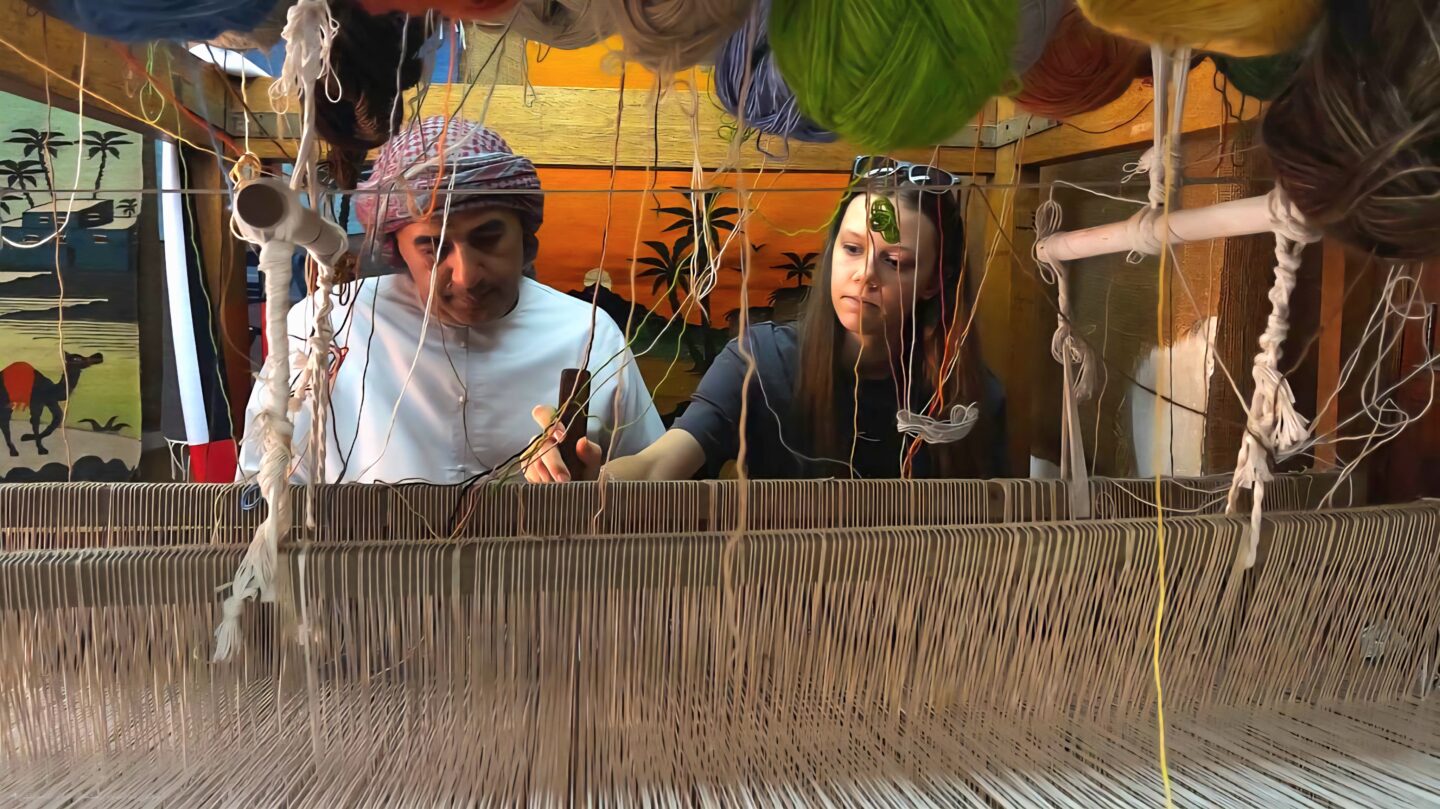 A man in traditional Emirati clothing teaches a woman how to weave on a wooden loom in Abu Dhabi. Colorful yarns hang above them, and a desert-themed tapestry with camels and palm trees decorates the background.